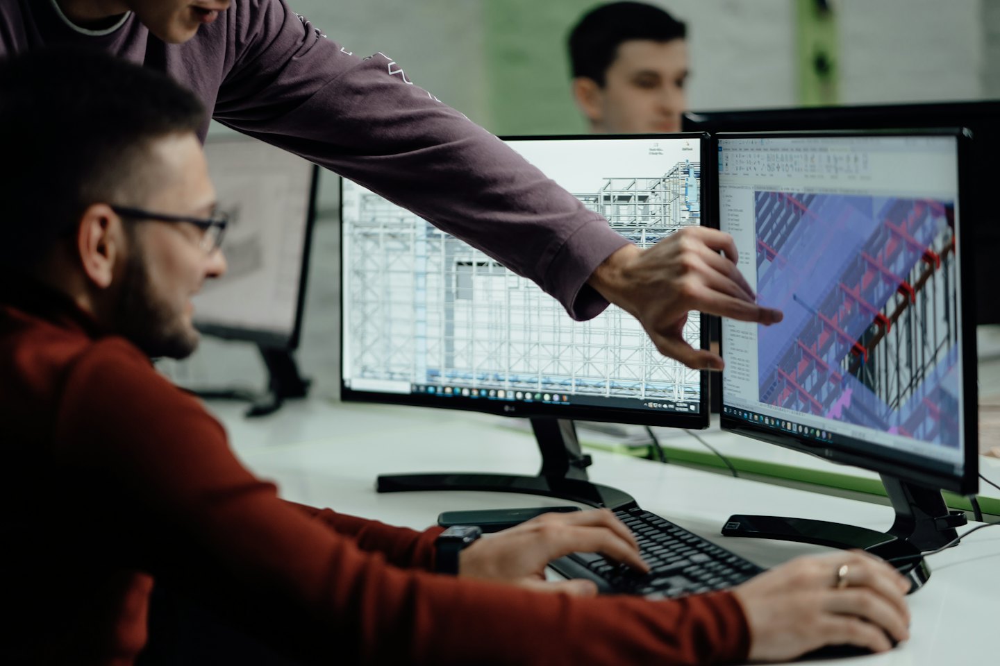 Hombre señala un diseño 3D en monitores duales mientras un colega escribe.