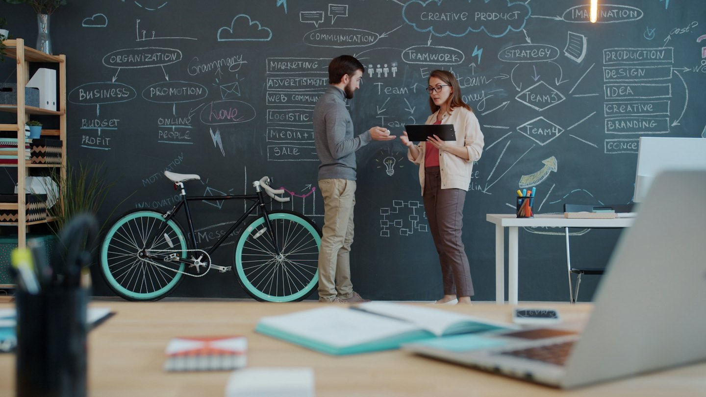 Dos profesionales discutiendo un plan de negocios con diagramas en una pizarra.